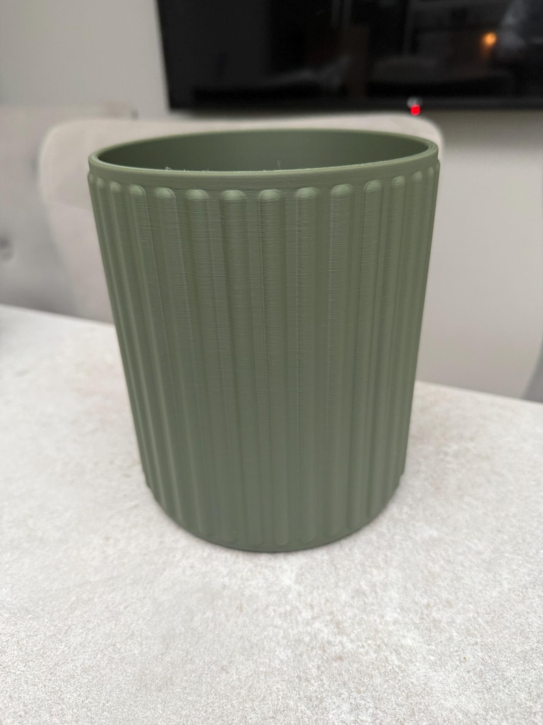 May include: A sage green cylindrical planter with vertical ribbed detailing. The planter has a slightly flared top and a smooth, rounded base. The color is a muted, earthy tone, suitable for various home decor styles. The planter is likely made of plastic or a similar material.