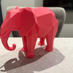 Op de afbeelding: Een koraalkleurige geometrische olifant sculptuur. De olifant heeft een low-poly ontwerp met veel platte oppervlakken. De slurf is gekruld en de poten zijn stevig. De sculptuur staat op een lichtgrijze ondergrond.