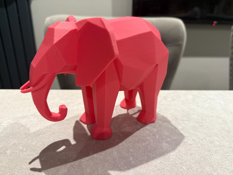 Op de afbeelding: Een koraalkleurige geometrische olifant sculptuur. De olifant heeft een low-poly ontwerp met veel platte oppervlakken. De slurf is gekruld en de poten zijn stevig. De sculptuur staat op een lichtgrijze ondergrond.