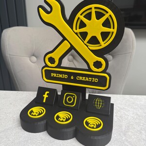 May include: A black and yellow 3D-printed display stand featuring a wrench, a wheel, and social media icons. The stand includes the text "PRIM3D & CREAT3D" and NFC symbols.