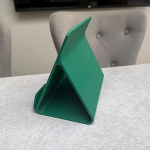 May include: A vibrant green tablet stand with a triangular design. The stand is made of a solid material and features a curved base and a raised back for device support. The stand is positioned on a light gray surface.
