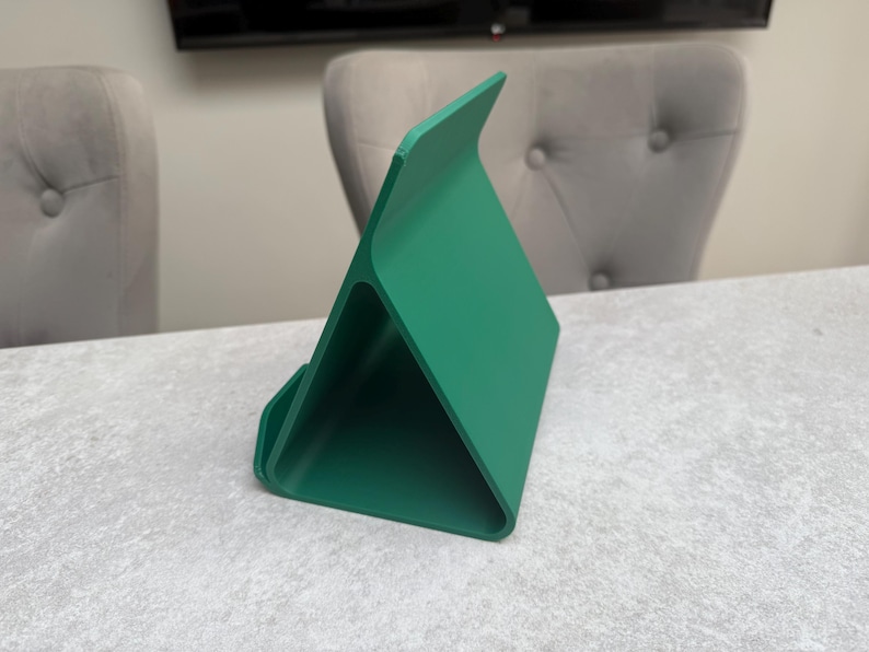 May include: A vibrant green tablet stand with a triangular design. The stand is made of a solid material and features a curved base and a raised back for device support. The stand is positioned on a light gray surface.