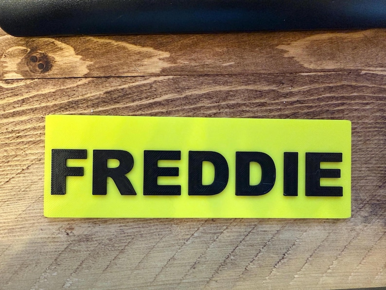 Pu&ograve; includere: Un'etichetta rettangolare con il nome "FREDDIE" in grassetto, lettere nere su uno sfondo giallo brillante. L'etichetta con il nome &egrave; posizionata su una superficie di legno con venature visibili.