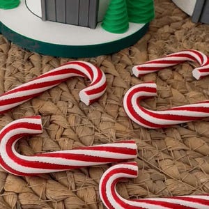 以下が含まれることがあります： 赤と白のストライプのキャンディケーン、小さな緑のクリスマスツリー、灰色のドアが付いた赤と白の家が特徴の、お祝いのクリスマスのシーン。アイテムは織りマットに配置されています。