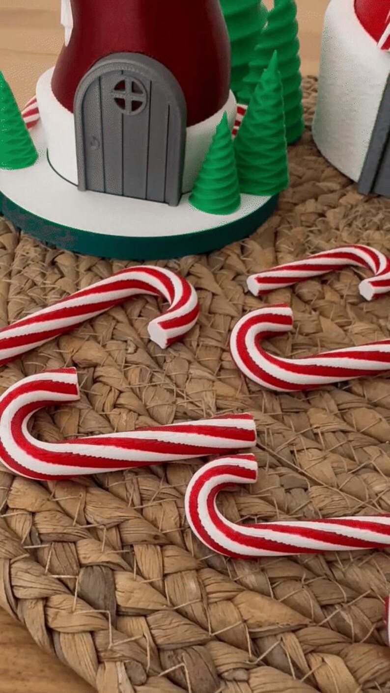 以下が含まれることがあります： 赤と白のストライプのキャンディケーン、小さな緑のクリスマスツリー、灰色のドアが付いた赤と白の家が特徴の、お祝いのクリスマスのシーン。アイテムは織りマットに配置されています。