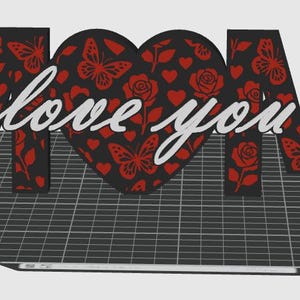 May include: A 3D-printed decorative piece spelling "MOM" with a heart-shaped "O". The letters and heart are black with red butterfly, rose, and heart patterns. The words "love you" are in cursive.