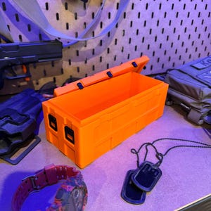 May include: Bright orange rectangular storage box with a hinged lid, open to reveal the interior. A red and black camouflage watch, black dog tags, and other tactical gear are also visible.