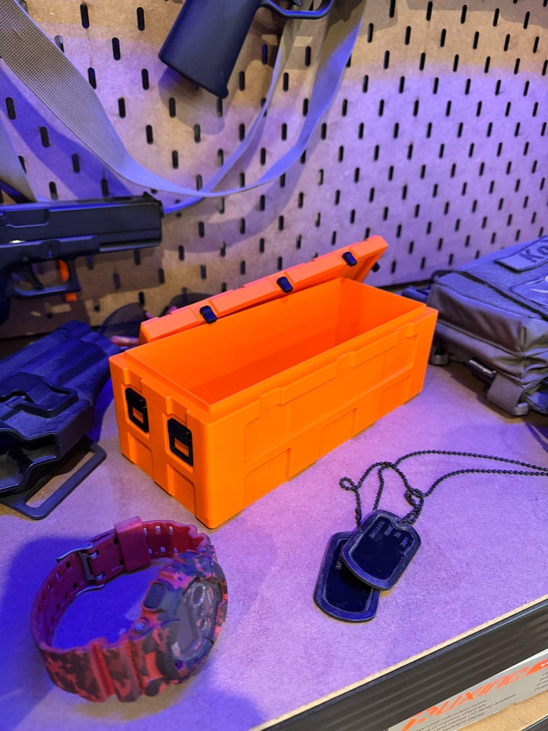May include: Bright orange rectangular storage box with a hinged lid, open to reveal the interior. A red and black camouflage watch, black dog tags, and other tactical gear are also visible.