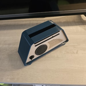 May include: A blue and beige retro-style speaker. The speaker has a rectangular shape with a slot on top and a circular speaker grill. The front panel is beige with a black speaker and a black button. The sides are blue.