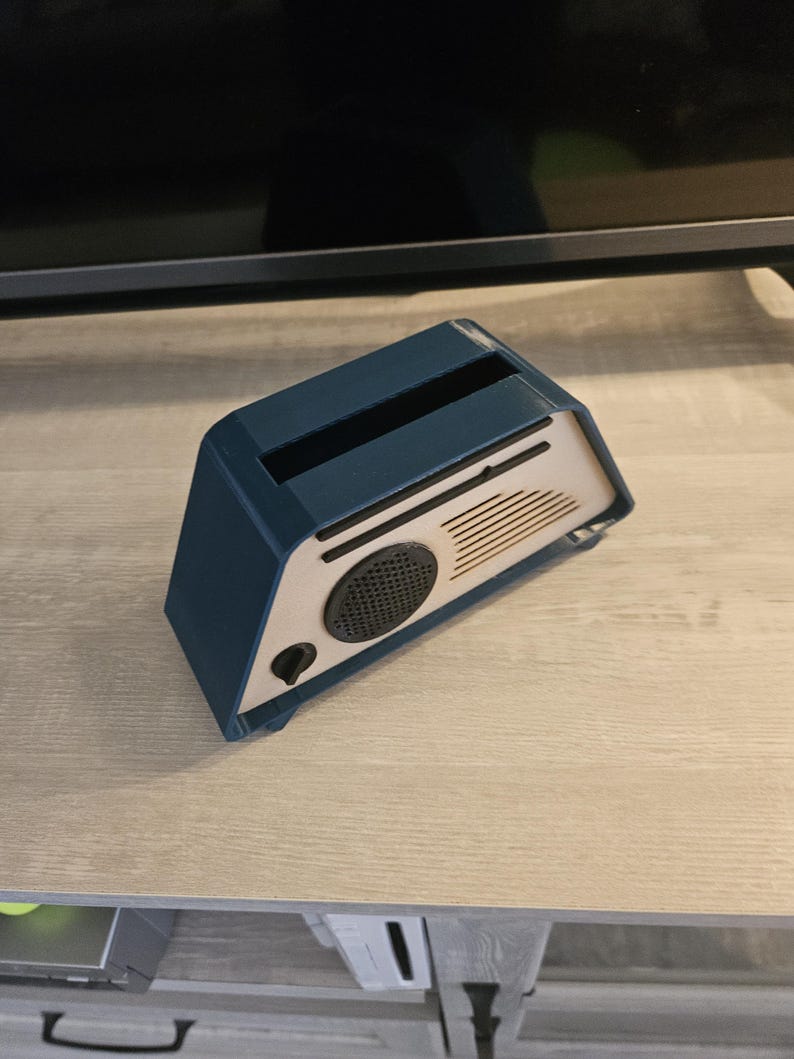 May include: A blue and beige retro-style speaker. The speaker has a rectangular shape with a slot on top and a circular speaker grill. The front panel is beige with a black speaker and a black button. The sides are blue.