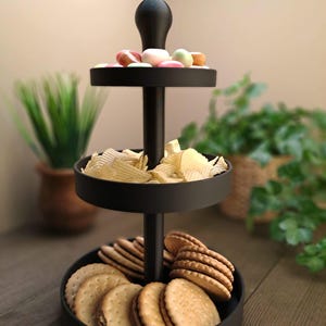 May include: A three-tiered black serving tray filled with snacks. The top tier holds colorful candies, the middle tier has potato chips, and the bottom tier is filled with cookies. The tray is on a wooden surface.