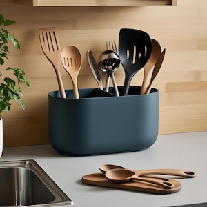 May include: A dark teal utensil holder containing wooden and black cooking tools. A wooden board with wooden spoons sits on the counter. Kitchen tools include a spatula, slotted spoon, ladle, and serving fork.