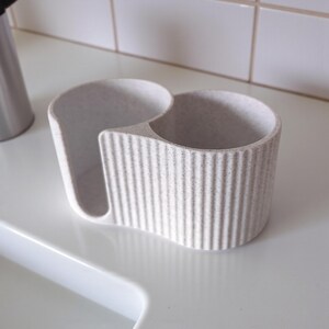 May include: A light gray kitchen sink caddy with a ribbed exterior. The caddy has two compartments, one for a sponge and the other for a brush or soap. The caddy is made of a speckled, textured material.