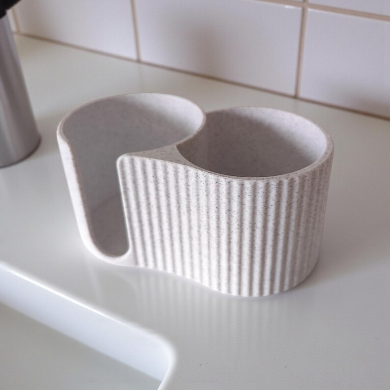 May include: A light gray kitchen sink caddy with a ribbed exterior. The caddy has two compartments, one for a sponge and the other for a brush or soap. The caddy is made of a speckled, textured material.
