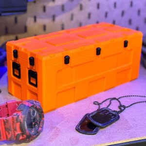 May include: An orange rectangular storage container with black latches and hinges. A red and black camouflage watch and a pair of black dog tags with a chain are in the foreground. The items are on a purple surface.