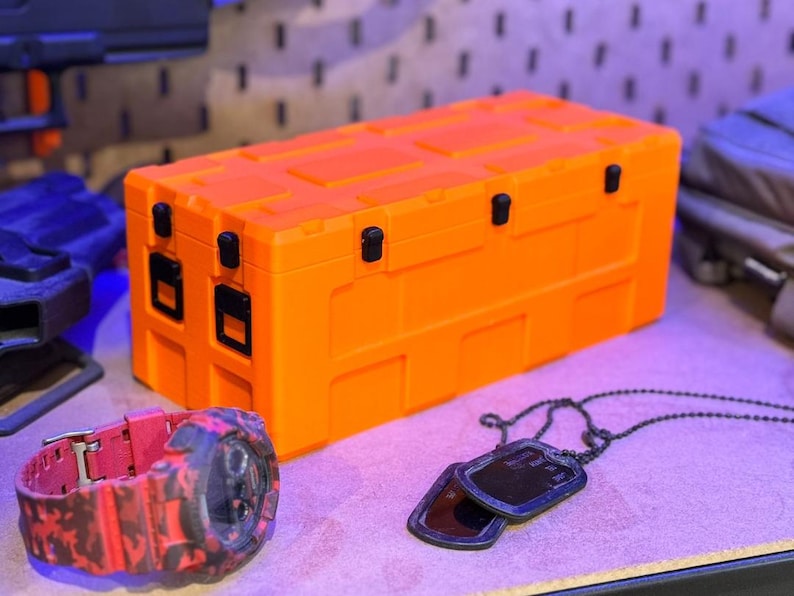 May include: An orange rectangular storage container with black latches and hinges. A red and black camouflage watch and a pair of black dog tags with a chain are in the foreground. The items are on a purple surface.