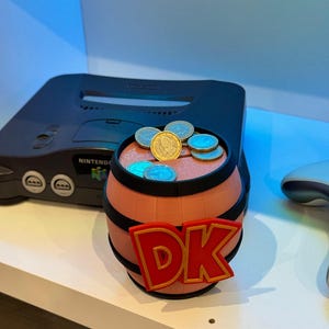 May include: A 3D-printed barrel with the letters "DK" in red and gold, filled with coins. The barrel is brown with black bands. A black Nintendo 64 console is in the background.