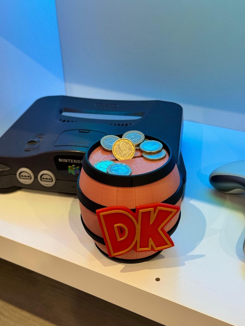 May include: A 3D-printed barrel with the letters "DK" in red and gold, filled with coins. The barrel is brown with black bands. A black Nintendo 64 console is in the background.