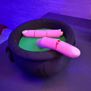 May include: A black cauldron with two pink finger-shaped objects inside. The cauldron sits on a green surface. One finger is segmented, and the other is cut open. The background is a dark purple.