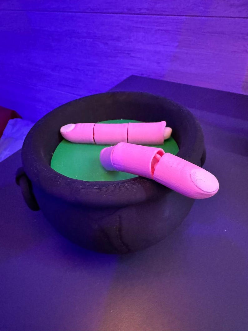 May include: A black cauldron with two pink finger-shaped objects inside. The cauldron sits on a green surface. One finger is segmented, and the other is cut open. The background is a dark purple.