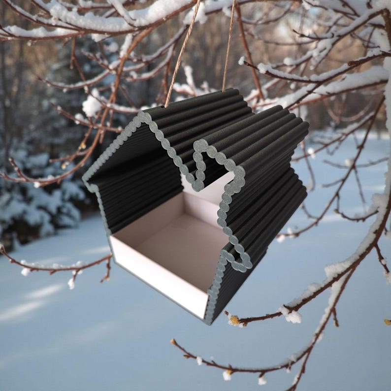 May include: A black and white birdhouse with a house-shaped design, hanging from a tree branch. The birdhouse has a corrugated roof and a white interior. The background shows a snowy landscape.