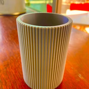 Op de afbeelding: Een cilindrische, beige container met verticale geribbelde details. De binnenkant is een donkerdere grijstint. De container staat op een roodbruin houten oppervlak. Het ontwerp suggereert een decoratief of functioneel item.