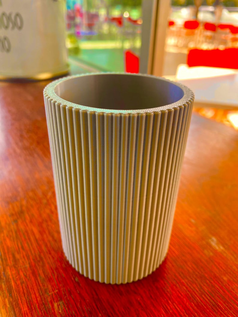 Op de afbeelding: Een cilindrische, beige container met verticale geribbelde details. De binnenkant is een donkerdere grijstint. De container staat op een roodbruin houten oppervlak. Het ontwerp suggereert een decoratief of functioneel item.