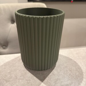 May include: A sage green cylindrical container with vertical ribbed detailing. The container is empty and sits on a light gray surface. The design is simple and modern, suitable for various uses.