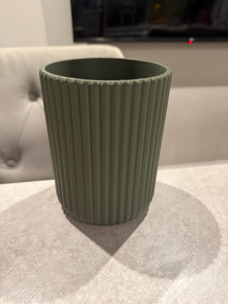 May include: A sage green cylindrical container with vertical ribbed detailing. The container is empty and sits on a light gray surface. The design is simple and modern, suitable for various uses.