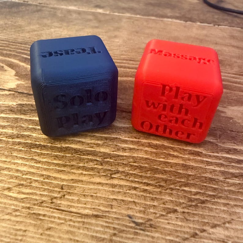Puede incluir: Dos dados impresos en 3D. Uno es azul con las palabras "Tease" y "Solo Play". El otro es rojo con "Massage" y "Play with each Other". Los dados est&aacute;n sobre una superficie de madera.