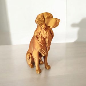 May include: A golden-colored dog figurine in a seated position. The sculpture has detailed fur texture and is posed looking to the side. The dog is sitting on a white surface.