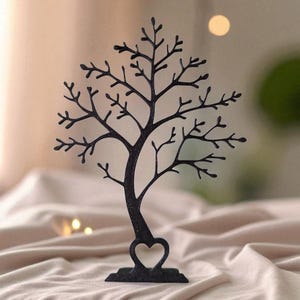 May include: Black metal tree-shaped jewelry holder with a heart-shaped cutout at the base. The tree has many branches and is designed to hold earrings, necklaces, and other accessories. The jewelry holder is displayed on a beige surface.