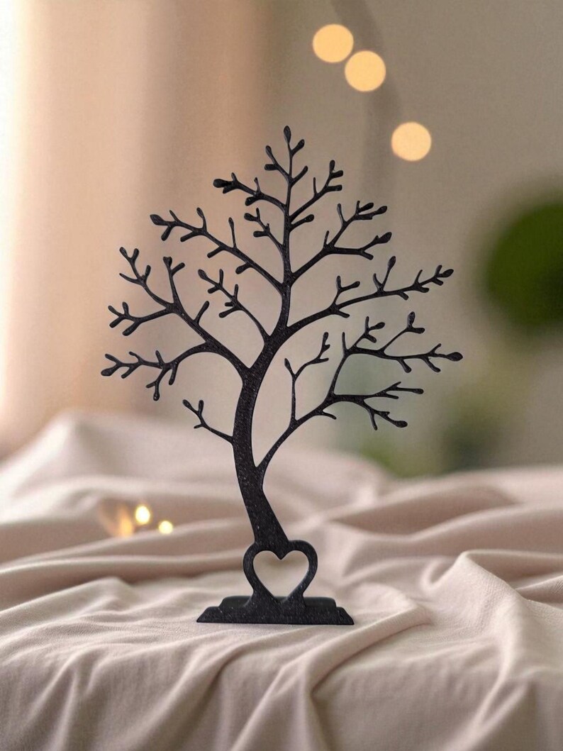 May include: Black metal tree-shaped jewelry holder with a heart-shaped cutout at the base. The tree has many branches and is designed to hold earrings, necklaces, and other accessories. The jewelry holder is displayed on a beige surface.
