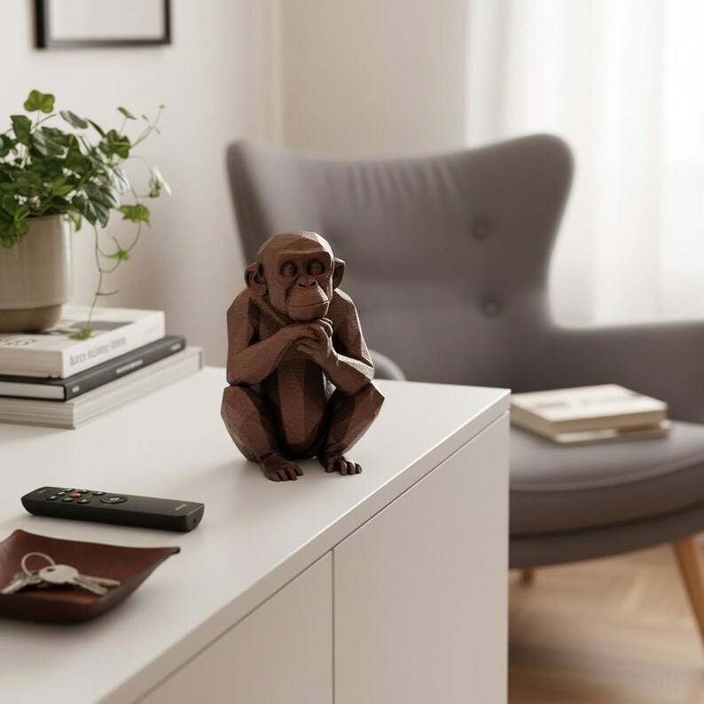 May include: A brown geometric chimpanzee sculpture sits on a white surface. The sculpture is a detailed representation of a chimpanzee in a seated position, with its hands clasped under its chin. The sculpture is a decorative item.