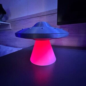 May include: A gray, 3D-printed UFO-shaped lamp with a red and pink glowing base. The lamp has a classic flying saucer design with small, raised details around the top. The lamp is on a dark surface.