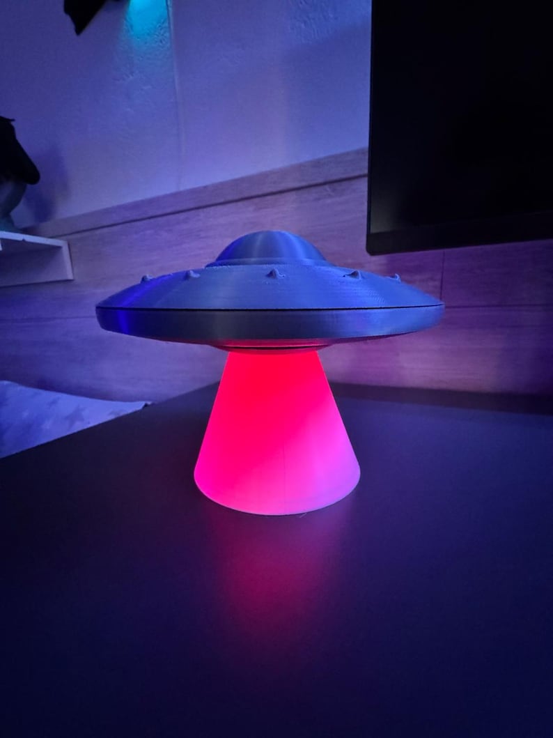 May include: A gray, 3D-printed UFO-shaped lamp with a red and pink glowing base. The lamp has a classic flying saucer design with small, raised details around the top. The lamp is on a dark surface.