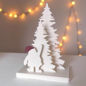 May include: A white Christmas-themed decoration featuring a stylized Santa Claus figure with a burgundy hat, standing in front of a white Christmas tree. The decoration is on a white base, with a string of warm-toned lights in the background.