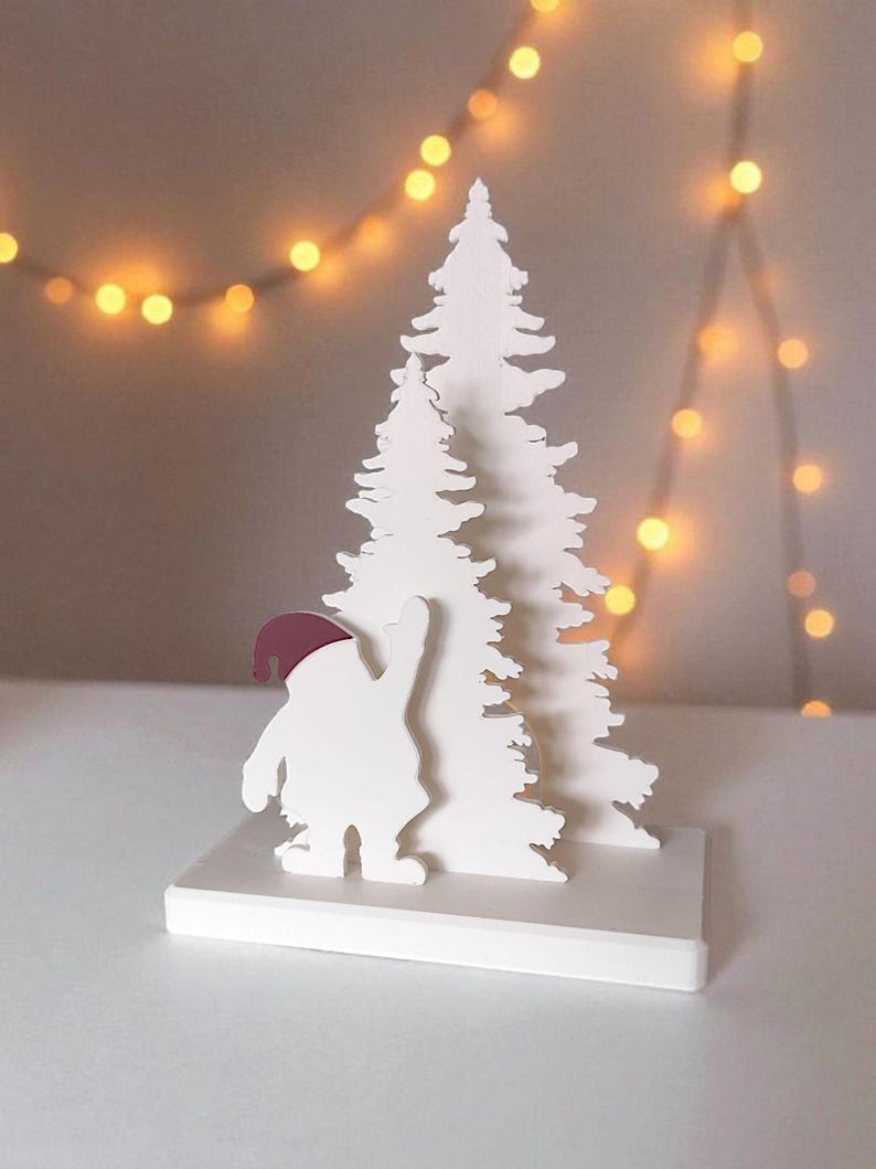May include: A white Christmas-themed decoration featuring a stylized Santa Claus figure with a burgundy hat, standing in front of a white Christmas tree. The decoration is on a white base, with a string of warm-toned lights in the background.
