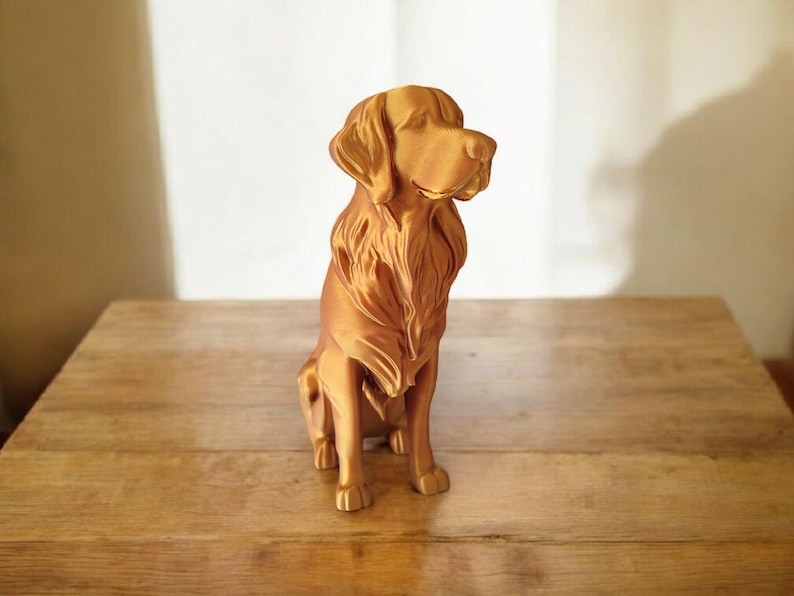 May include: A golden-colored dog statue, likely a Golden Retriever, sits on a wooden surface. The sculpture has detailed fur and a focused gaze, showcasing the breed's characteristics. The statue is a decorative item.