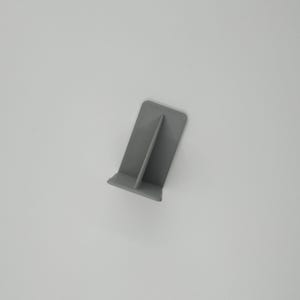 Op de afbeelding: Een kleine, grijze, plastic telefoonstandaard. De standaard heeft een rechthoekige basis met een verticaal steundeel. De standaard is ontworpen om een telefoon rechtop te houden. De standaard is gemaakt van effen, matte grijze kunststof.
