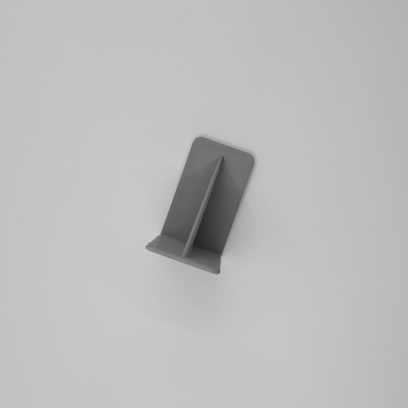 Op de afbeelding: Een kleine, grijze, plastic telefoonstandaard. De standaard heeft een rechthoekige basis met een verticaal steundeel. De standaard is ontworpen om een telefoon rechtop te houden. De standaard is gemaakt van effen, matte grijze kunststof.