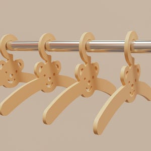 Pu&ograve; includere: Quattro grucce di legno per bambini con un design a forma di orsetto. Le grucce sono di colore beige chiaro e sono appese a un'asta di metallo argentato. Lo sfondo &egrave; di colore beige chiaro.