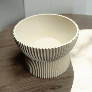 May include: A cream-colored, ribbed bowl with a scalloped rim sits on a pedestal base. The bowl's interior is smooth, contrasting with the textured exterior. The design is modern and minimalist, suitable for various home decor styles.