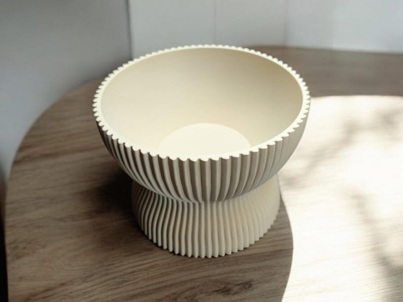 May include: A cream-colored, ribbed bowl with a scalloped rim sits on a pedestal base. The bowl's interior is smooth, contrasting with the textured exterior. The design is modern and minimalist, suitable for various home decor styles.