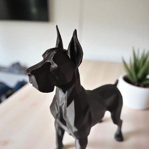 Puede incluir: Una estatua de perro Gran Dan&eacute;s negro, de estilo geom&eacute;trico. La escultura tiene un dise&ntilde;o facetado, con orejas puntiagudas y una cola corta. El perro est&aacute; sobre una superficie clara, con una planta en maceta en el fondo.
