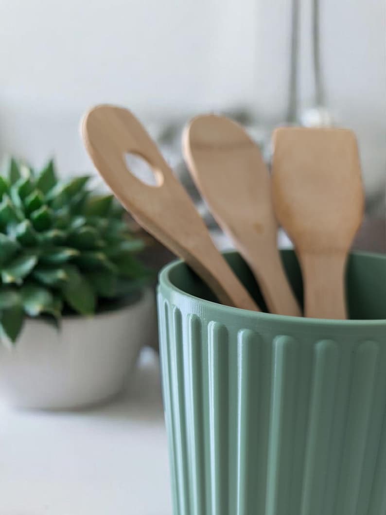 May include: A light green, ribbed ceramic utensil holder with three wooden cooking spoons. One spoon has a hole, another is solid, and the third is a spatula. A small potted succulent plant is in the background.