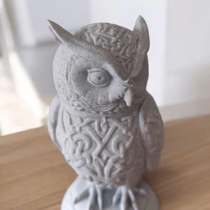 May include: A gray owl figurine with detailed feather and wing patterns. The owl has large eyes and a small beak. The figurine is made of a stone-like material and sits on a round base. The owl's chest features an intricate design.