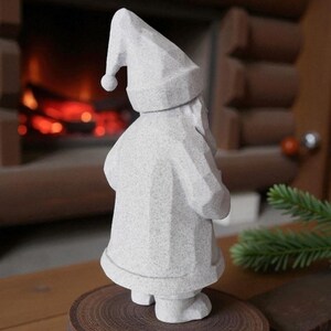 K&ouml;nnte beinhalten: Eine graue, geometrische Weihnachtsmannfigur mit spitzem Hut und Mantel, die auf einer Holzscheibe steht. Die Figur hat eine strukturierte Oberfl&auml;che und steht vor einem Kamin.