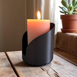 May include: A burning peach-coloured pillar candle in a black metal holder. The candle is centrally positioned, with a flame at the top. The holder has a curved design. The background includes a wooden surface and a potted plant.