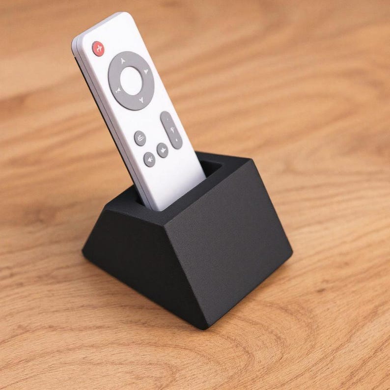 May include: A white remote control with gray buttons and a red power button is placed in a black, geometric holder. The holder is a truncated pyramid shape, and the remote is angled slightly. The background is a wooden surface.